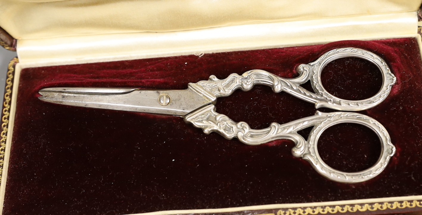 A cased set of twelve early 20th century French white metal fancy pattern teaspoons, a similar cased preserve spoon and a similar cased pair of white metal handled grape shears.